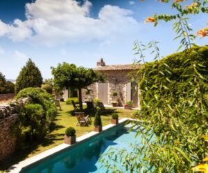 Résidence secondaire en France - Design d'Espaces - Vue d'une maison de vacances en pierre, avec volets, depuis le jardin avec piscine et luxurieuse végétation